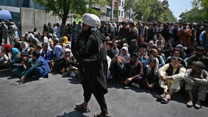 A Taliban fighter walks in front of people sitting along a road outside a bank waiting to withdraw money in Kabul, on 4 September, 2021 | Photographer: Wakil Kohsar/AFP/Getty Images via Bloomberg